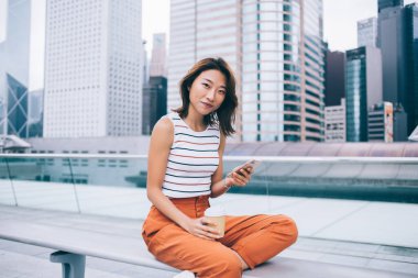 Güzel genç Asyalı bir kadın gülümsüyor ve bacak bacak üstüne atmış bir şekilde bankta oturuyor. Plastik bardak ve akıllı telefon ile Hong Kong şehrinin arka planındaki kameraya bakıyor.