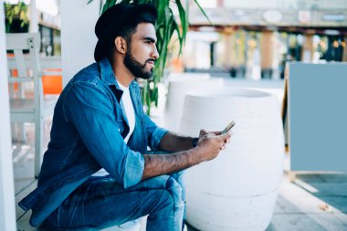 İspanyol rüyacı, barışçıl, kot elbiseli, şık şapkalı, akıllı telefonlu, şehir kafesinin merdivenlerinde dinlenen ve gözlerini kaçıran adamın yan görüntüsü.