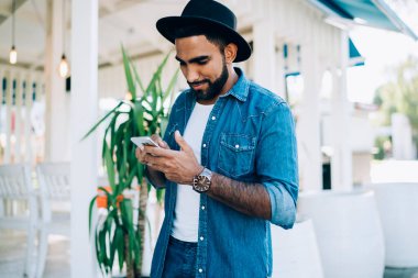 İyimser sakallı, modaya uygun kıyafetli, şapkalı, gülümseyen ve akıllı telefon kullanan bir etnik adam güneşli bir günde güzel bir kafe önünde dikiliyor.