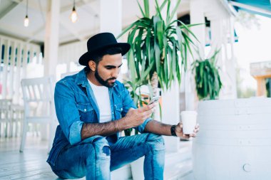 Kot pantolon ve yaz şapkası giyen düşünceli İspanyol erkek dinlenme saatlerinde teras basamaklarında kahve eşliğinde dikkatli bir şekilde sörf yapıyor.