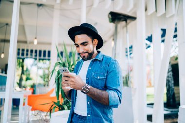 Siyah şapkalı, sıradan giysili, siyah şapkalı, düşük açılı mutlu İspanyol bir erkek. Ekrana odaklanıyor ve modern kafenin bulanık iç kısımlarına karşı durarak akıllı telefonla iletişim kuruyor.