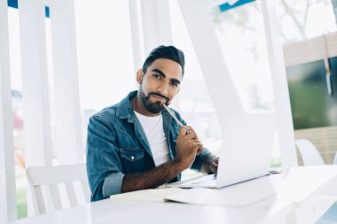 Gündelik giysiler içinde, masada oturmuş cafe 'nin şık açık hava kameralarına bakarken dizüstü bilgisayarla çalışmakla meşgul etnik bir erkek serbest çalışıyor. 
