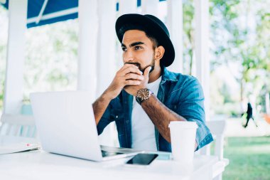 Düşük açılı, düşünceli, siyah şapkalı, sıradan kıyafetleri olan İspanyol bir adam beyaz masaya oturmuş, elinde kahve ve modern kafede netbook kullanırken kafasını çeviriyor.