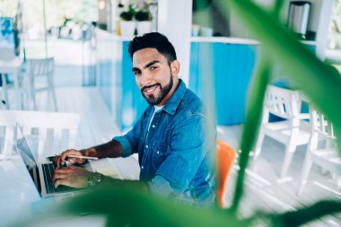 Modern kafede dizüstü bilgisayarda sörf yaparken gülümseyen ve kameraya bakan kendine güvenen etnik erkek girişimcinin yan görüntüsü 