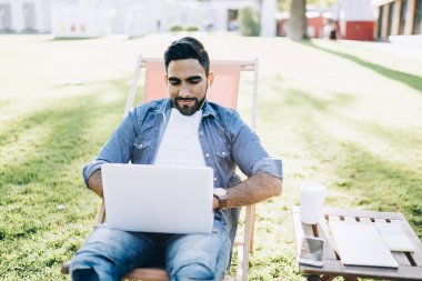 Normal kot gömlekli ve kot pantolonlu yakışıklı etnik erkek yazın parkta, dinlenme odasında otururken dizüstü bilgisayarını karıştırıyor.