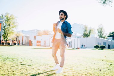 Sıradan kot gömlekli ve siyah şapkalı yırtık pantolonlu, yazın parkta yürüyen zeki, etnik bir erkek.