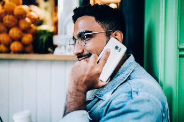 Gözlüklü ve kot ceketli neşeli etnik erkeğin telefon konuşması yapması ve meyve dükkanının yanında otururken mutlu bir şekilde gülümsemesi. 