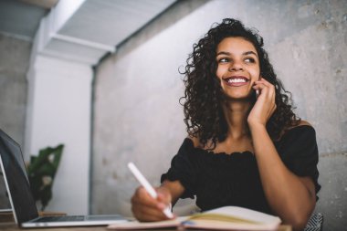 Düşük açılı neşeli kıvırcık saçlı İspanyol kadın. Günlük giysiler içinde deftere yazıyor ve cep telefonunu ararken dizüstü bilgisayar kullanıyor.