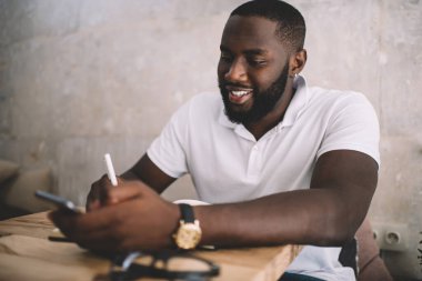 Beyaz tişörtlü neşeli siyah sakallı adam not alıyor ve kafede otururken akıllı telefon kullanıyor.