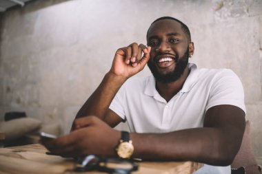 Cep telefonuyla mesajlaşırken ve kafede oturmuş kameraya bakan sıradan giyinmiş neşeli, Afro-Amerikalı genç bir adam.