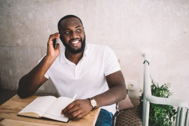 Modern kafede oturmuş, cep telefonuyla konuşan ve notlar alan gündelik kıyafetli mutlu siyah adam.