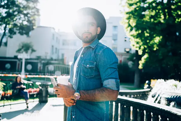 Low angle in sunbeams of stylish thoughtful Hispanic businessman in hat and casual clothes with takeaway coffee and smartphone looking away and thinking about future while standing on street