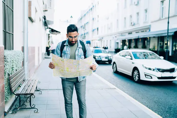 Confused puzzled bearded ethnic male in glasses in casual denim jacket standing in street holding map in hand in big city