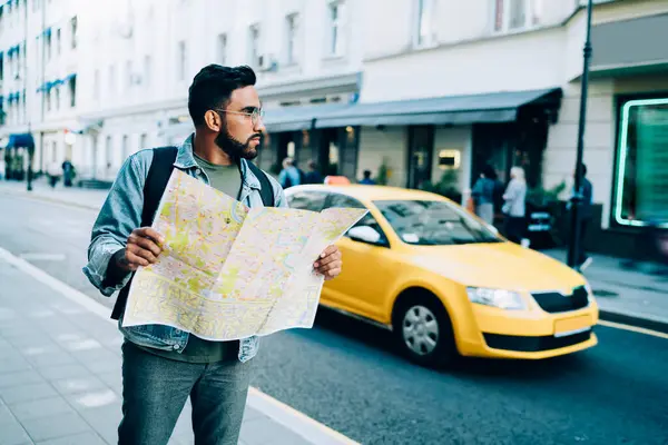 Puzzled  curious bearded ethnic male in glasses in casual denim jacket standing in street holding map in hand in big city
