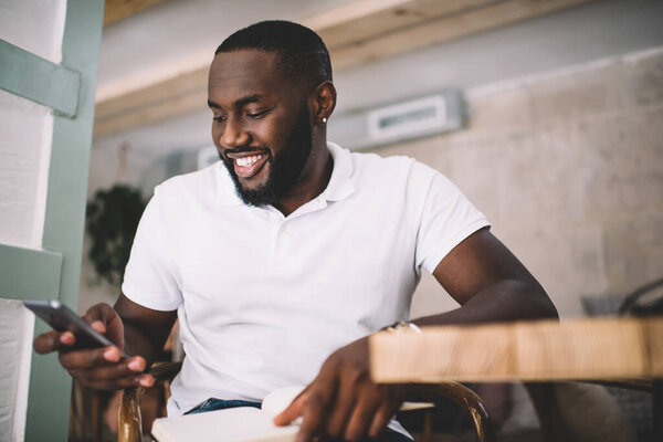 Cheerful young African American man in casual clothes using cellphone while holding notebook and sitting at table in modern cafe