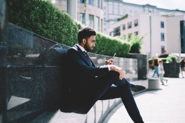 Resmi olarak giyinmiş sakallı bir adamın elinde cep telefonuyla bankta oturması ve güneşli bir günde iş merkezinin yanında mesajlaşması.