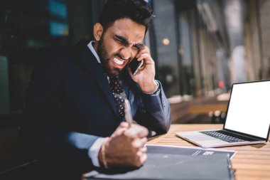 Sinirli iş adamı, siyah takım elbiseli, beyaz gömlekli, ağ defteri takan, cep telefonuyla şehirdeki kafede ahşap masaya yumruk atan kurşunu ısırıyor. 