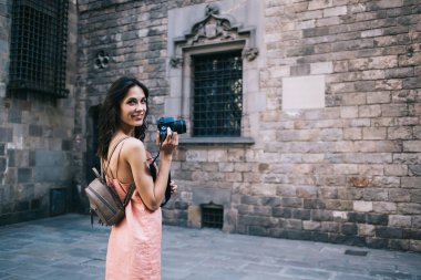 Yan görüntüde, şeftali rengi elbiseli, dijital kameralı çekici bir kadın eski kasabadaki taş devrinden kalma binaya karşı caddede dikiliyor.