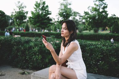 Beyaz elbiseli şık esmer kadının yan görünüşü. Dikkatlice sörf yapan cep telefonu. Sokakta yeşil çitli bankta oturuyor. 