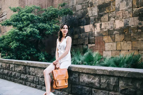 Deri sırt çantalı mutlu bir kadın yaşlı taş çitlerin üzerinde oturmuş bahçe bitkilerine karşı yazın eski kasabayı keşfediyor ve kameraya gülümsüyor.