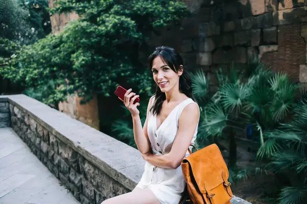Siyah saçlı, beyaz elbiseli, sırt çantası ve cep telefonuyla yaz günü şehirde oturan mutlu bir gezgin kadın.