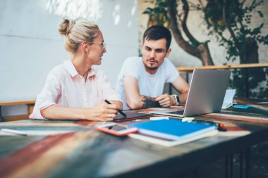 Kalem ve tabletle genç sarışın kadın, kafenin terasında dizüstü bilgisayarın önünde otururken günlük çalışma sürecini erkek meslektaşına anlatıyor.