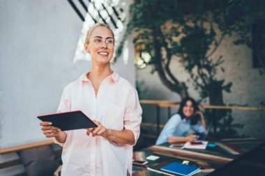 Keyifli bir kadın tablet üzerindeki verileri tarıyor ve masaların yanında durup terasta bulanık bir şekilde gülümsüyor.