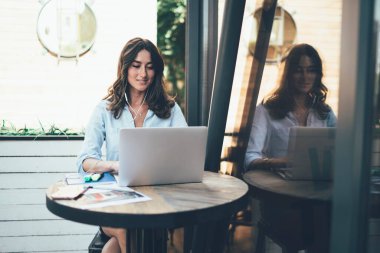 Uzun siyah saçlı, gülümseyen iş kadını müzik dinliyor ve laptopunu yuvarlak masada oturuyor ve dışarıdaki kafenin camına yansıyor.