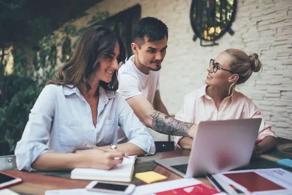 stock image Group of cheerful coworkers standing and sitting at desk outdoors looking at screen of laptop, talking and writing in planner