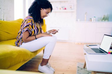 Masanın yanında dizüstü bilgisayarı boş bir şekilde otururken şık giyinmiş, gülümseyen ve akıllı telefon kullanan neşeli siyahi gencin yan görüntüsü.