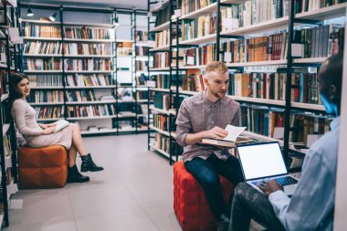 Kitap okuyan genç bir adam, kütüphanede kitaplıkların ve bayan sınıf arkadaşının yanında oturup ödev yaparken siyahi arkadaşına bakıyor.