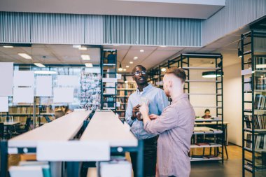 Üniversite kütüphanesinde kitaplığın yanında durmuş ders kitabından veri tartışan ve birlikte okuyan genç çoklu etnik erkeklerin yan bakışı