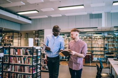 Genç, çok ırklı erkek öğrenciler gündelik kıyafetlerle edebiyatı tartışırken, elinde kitap tutarken ve kütüphanede konuşurken. 