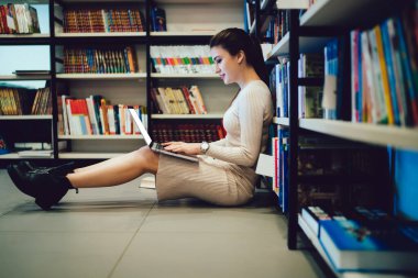 Kendine güvenen, çalışkan bir öğrenci kadının kitap dolu bir kütüphanede yerde oturup kitaplığa yaslanmasının yan görüntüsü.