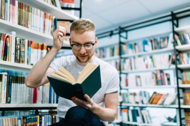 Düşünceli yetişkin sakallı adam gündelik giysiler ve gözlüklerle kitaba odaklanıyor ve aydınlık modern kütüphanede el kaldırıyor.