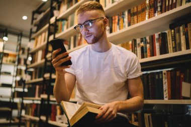 Gündelik giysiler ve gözlük takmış, dizleri açık ve modern kütüphanede kitaplıkların yanında akıllı telefon kullanan dalgın, yetişkin sakallı bir öğrenciden.