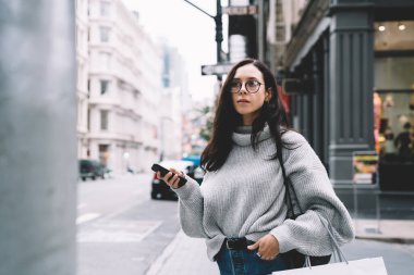 Gözlüklü, sırt çantalı, alışveriş çantalı ve akıllı telefonlu modern bir kadın New York 'a bakıyor.