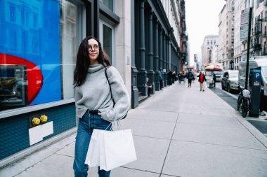 Neşeli, gevşemiş, kazaklı, kot pantolonlu bir kadın alışveriş torbalarıyla kaldırımda yürüyor ve New York 'ta hayallere dalmış gibi bakıyor.