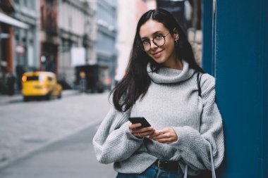 Güzel, genç, güzel, gülümseyen esmer, şık gözlüklü, gri kazaklı, akıllı telefonu elinde tutarak şehir sokaklarında kameraya bakıyor.