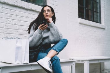 Çekici, ince, düşünceli, gözlüklü, akıllı telefon kullanarak kafasını çeviren, alışveriş poşetlerinin yanında bankta oturan...