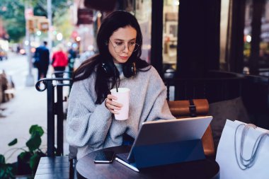 Gri kazaklı, gözlüklü, kulaklık takan ve New York 'taki kafede dijital tablet üzerinde çalışırken içki içen güzel bir kadın. 