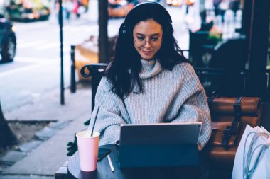 Süveterli, gözlüklü, kulaklık takan ve tablet kullanan genç ve güzel bir esmer. Caddedeki kafede içki içiyor.