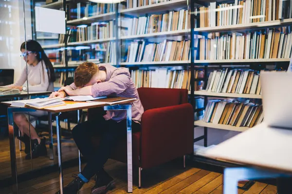 Tired male employee dressed in formal suit lying face down on desk with books and notepads in library near bookshelves