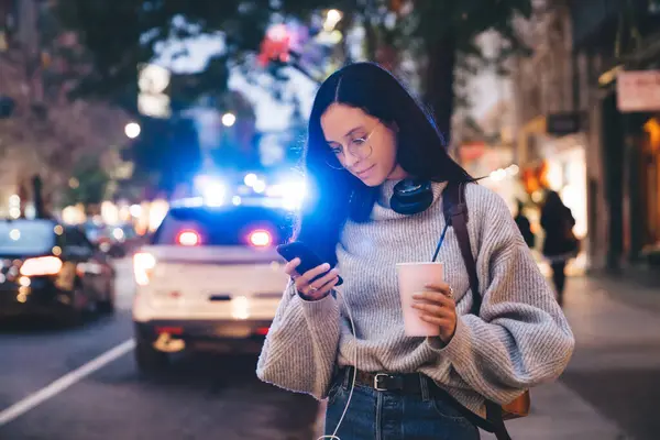 Attractive casual modern woman in glasses and headphones having drink to go and using modern smartphone on busy street in evening