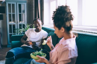 Çok ırklı, neşeli genç kadın ve erkek, gündelik giysiler içinde hafif bir kanepede yan yana otururken yiyip içiyorlar.