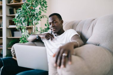 Beyaz tişörtlü düşünceli Afro-Amerikalı adam evde takı takıyor, dizüstü bilgisayar kullanıyor ve gündüz vakti aşağı bakıyor.