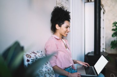 Gündüz vakti rahat bir odada otururken rahat bir şekilde dizüstü bilgisayar kullanan Afro-Amerikan kadınlarının yan görüntüsü.