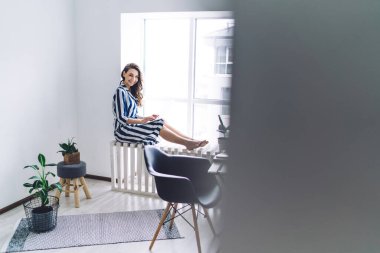 Çağdaş İskandinav dizayn dairesinde çizgili uzun tişörtlü, mutlu genç bir kadın pencerenin yanında ev bitkileriyle oturuyor.