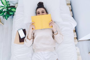 Beyaz keten ve akıllı telefonlu yatakta uzanırken gülen gözlü, yarı yüzünü açık sarı kitapla kapatan büyüleyici bir kadından. 