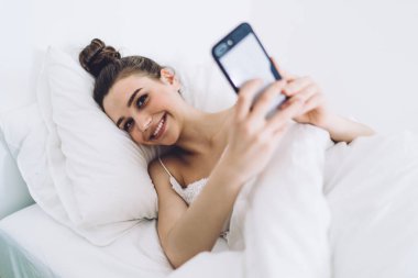 Huzurlu esmer, beyaz yastıklı, rahat bir yatakta uzanmış selfie çekerken beyaz arka plandaki odada kameraya bakıyor.